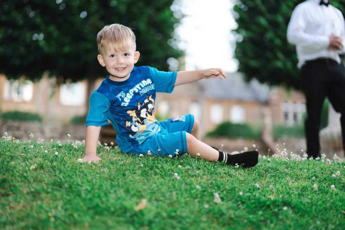 A young boy happily sits and smiles on a lush green lawn outdoors during a wedding reception celebration.