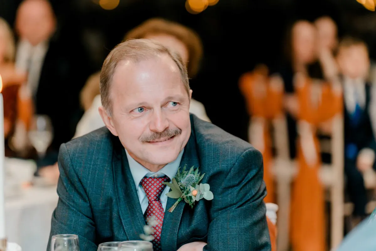 A mature male wedding guest wearing a checked suit and patterned tie with a thistle buttonhole smiles candidly during the wedding reception.