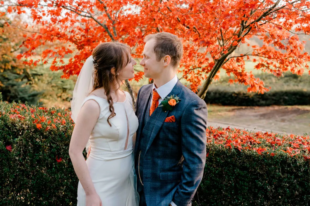A bride and groom share a tender moment face-to-face outdoors against a vibrant backdrop of autumn leaves and brilliant red foliage.
