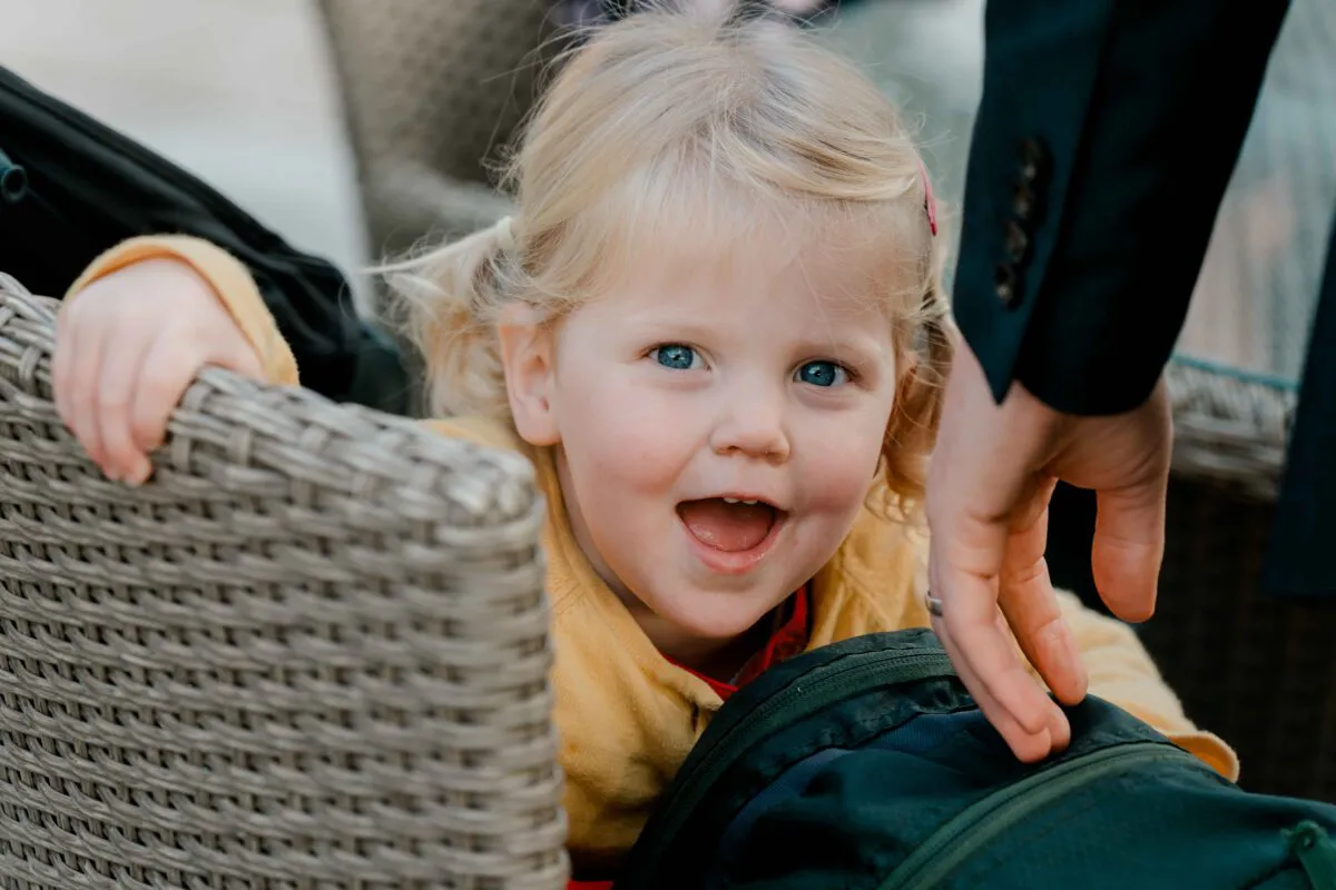 A young blonde flower girl laughs happily while peeking over a rattan chair during the wedding celebration.