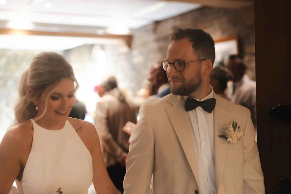 A newlywed couple shares a warm glance as they walk hand in hand through an indoor venue during their wedding celebration.