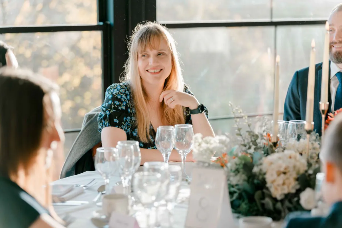 A smiling guest with blonde hair and a floral top sits at a wedding reception table set with candles and floral centrepieces.
