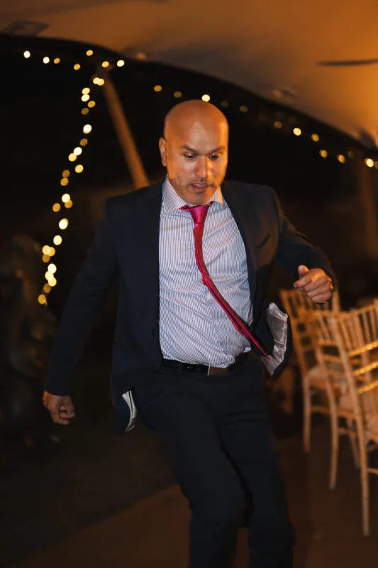 A guest in a navy suit and bright pink tie enthusiastically dances at an evening reception illuminated by fairy lights.