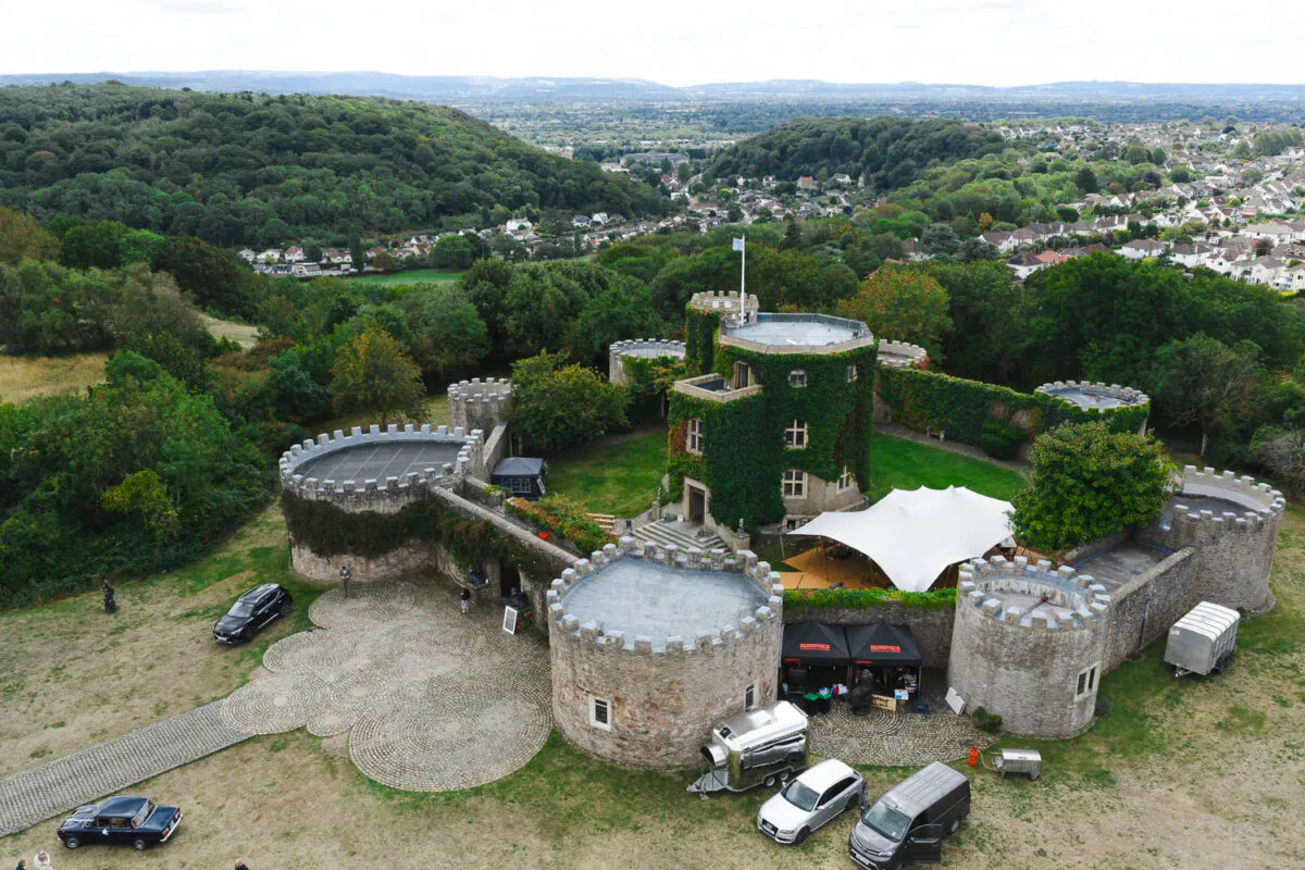 An aerial drone shot showcases a historic stone castle venue surrounded by lush green forests and a distant town, with a white stretch tent set up in the courtyard.