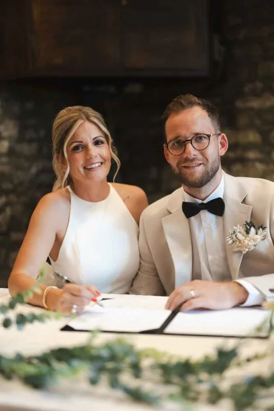 A smiling bride and groom sit side by side signing their wedding register, with the bride wearing a sleek halter-neck dress and the groom wearing a beige suit and black bow tie.