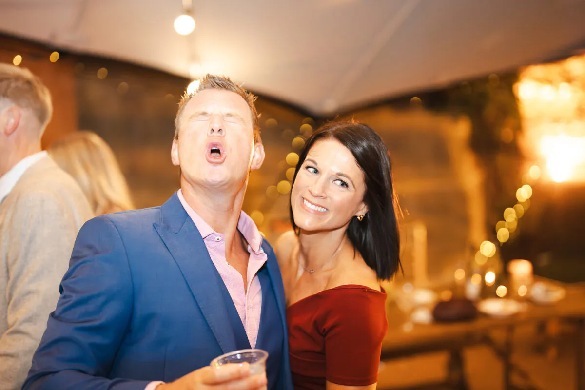 A guest in a blue suit makes a funny face while holding a drink next to a smiling woman at an evening wedding reception.