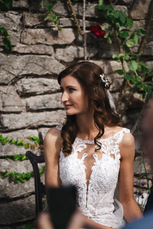 A smiling bride with wavy brown hair wearing an intricate lace dress with a plunging illusion neckline, seated outdoors against a rustic stone wall.