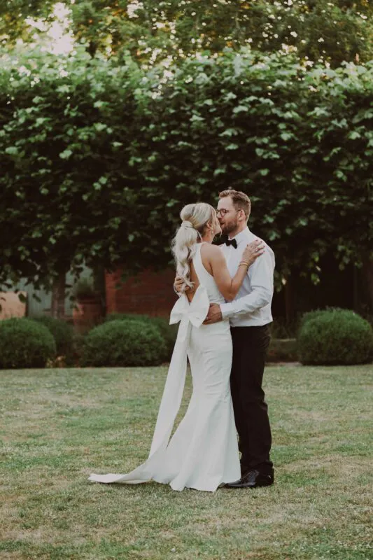 A newlywed couple shares a tender kiss in a lush green garden, with the bride wearing a sleek halter-neck gown featuring an oversized bow detail on the back.