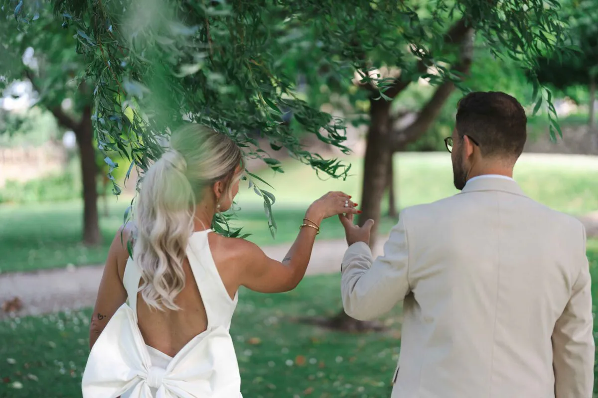 A newlywed couple is captured from behind walking hand in hand through a lush green garden underneath hanging tree branches on their wedding day.