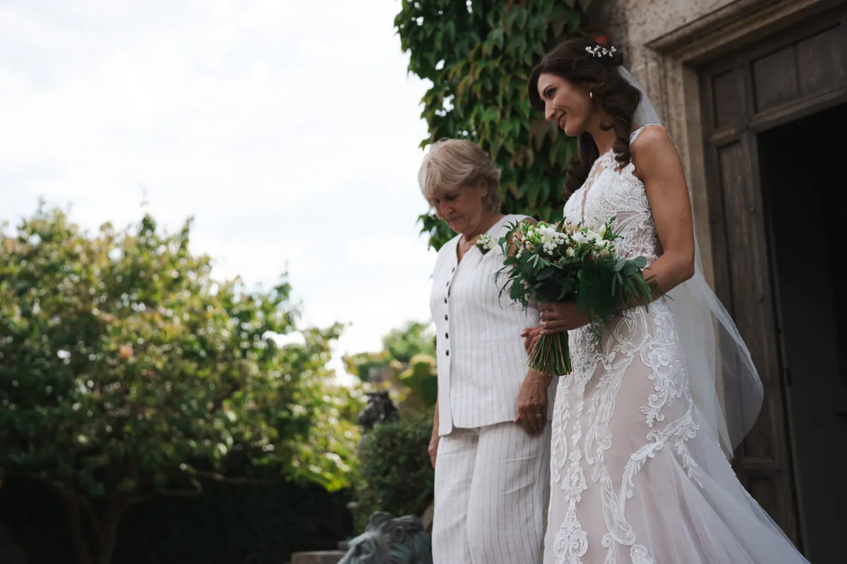 A smiling bride holding a hand-tied bouquet walks alongside her mother outside near a stone doorway framed by ivy.