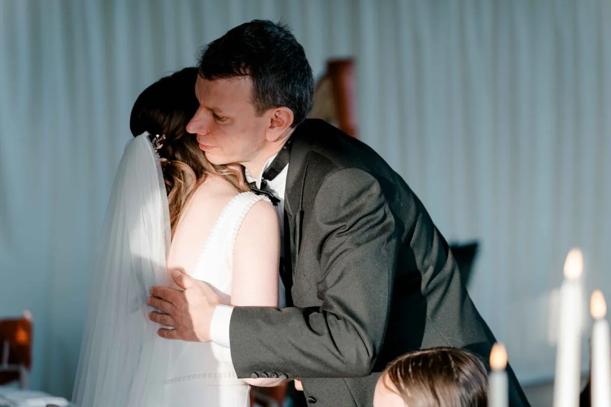A groom in a classic black tuxedo warmly embraces his bride, who wears a sleeveless white dress and veil, bathed in soft side lighting.