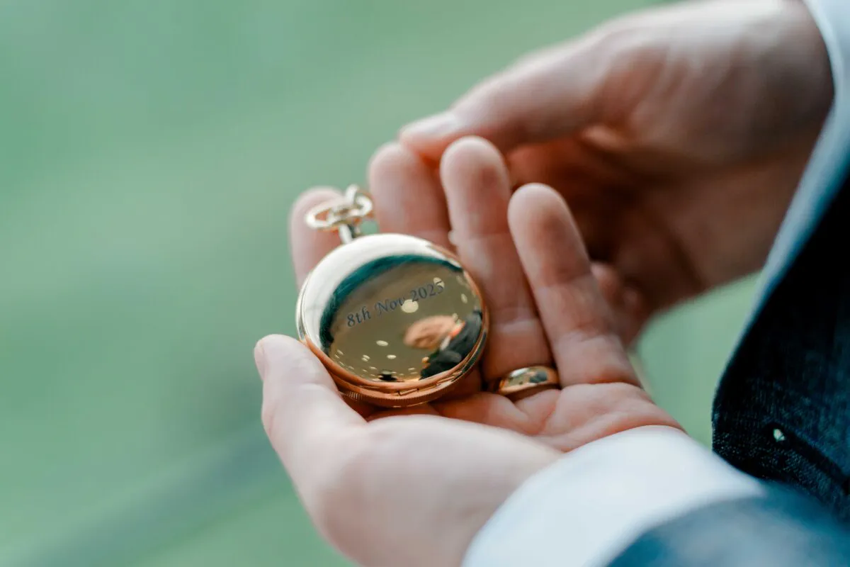 Close-up of a groom holding a polished gold pocket watch engraved with a wedding date during morning preparations.