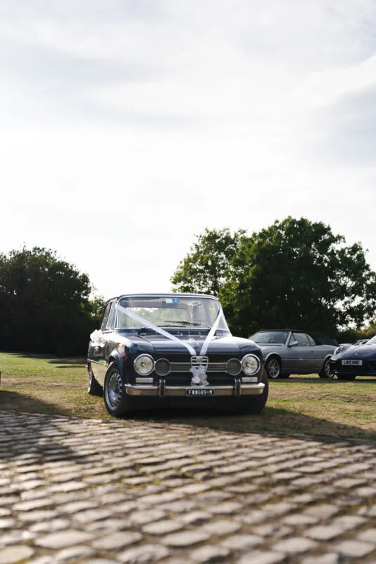 A vintage dark wedding car decorated with white ribbons is parked on a cobbled courtyard outdoors under a bright sky.