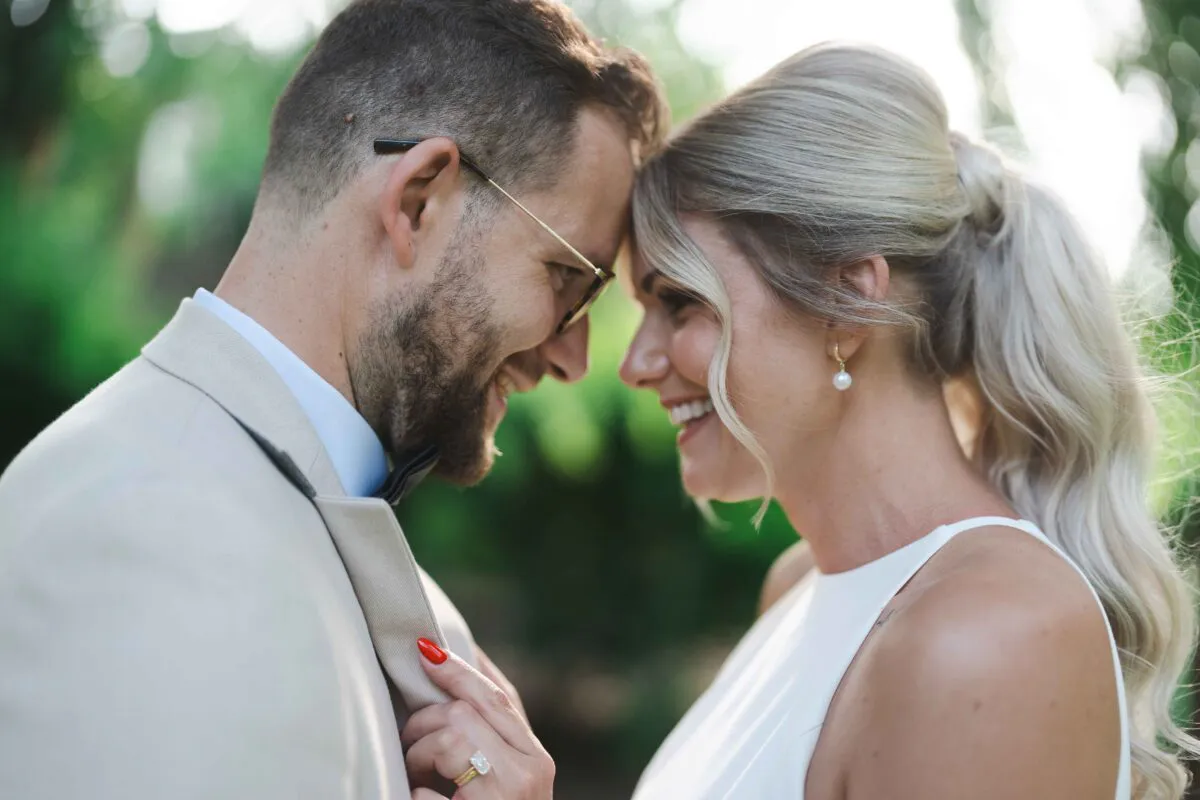 A smiling bride and groom share a tender nose-to-nose moment outdoors, with the groom wearing a beige suit and glasses and the bride sporting a high ponytail and pearl earrings.