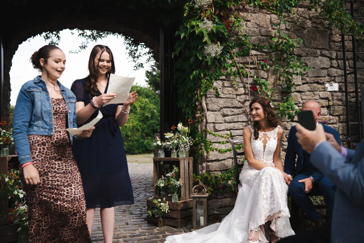Two wedding guests laugh while reading speeches near an ivy-covered stone wall, while the bride looks on smiling from a seated position.