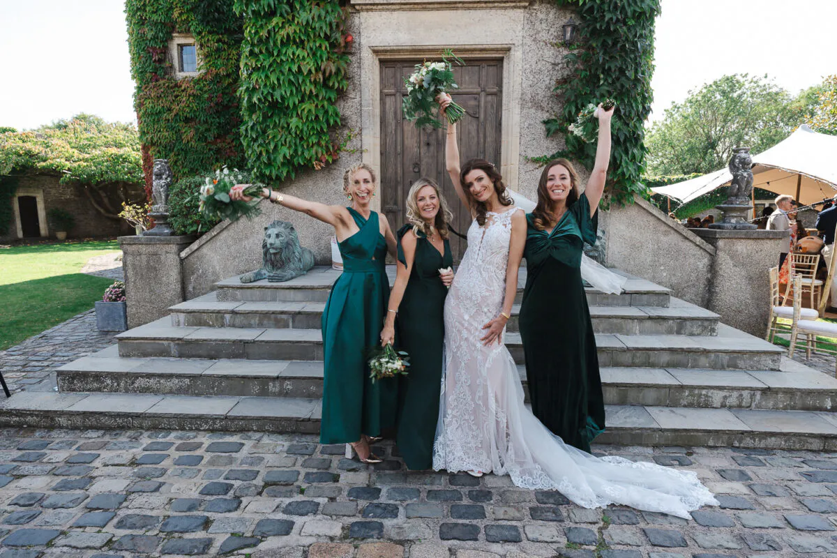 A smiling bride in a fitted lace gown with a dramatic train poses alongside three bridesmaids wearing deep green velvet dresses, all raising their bouquets in celebration outside an ivy-clad stone building.