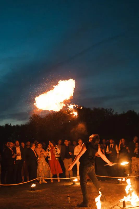 A fire breather performs an impressive flame display at an outdoor evening wedding reception while guests watch in awe under a deep blue twilight sky.