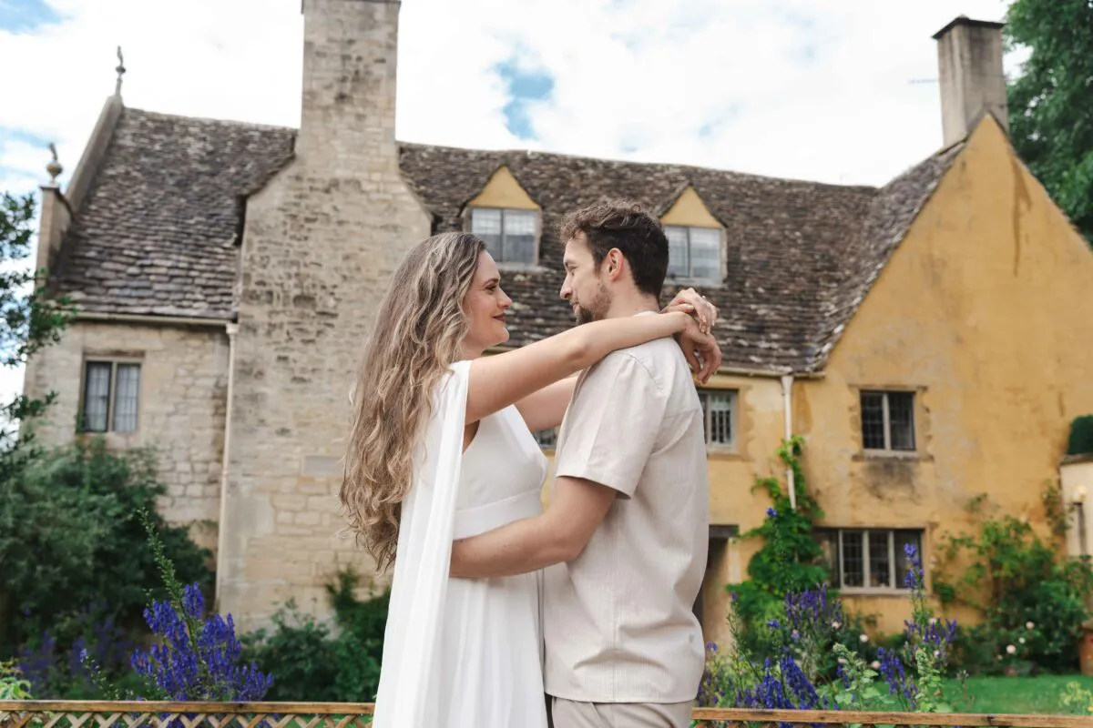 A newlywed couple embraces lovingly outdoors with a historic stone manor house and vibrant garden flowers in the background.