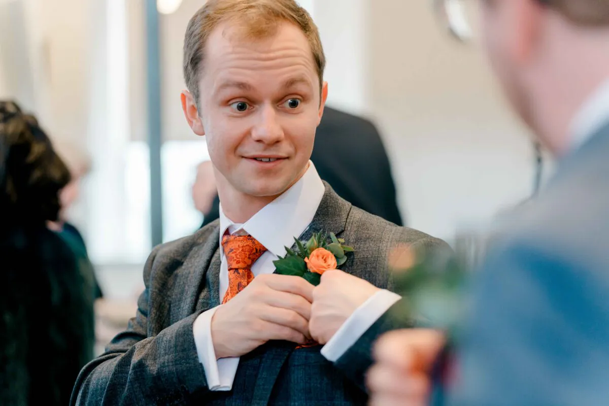 A groom in a grey suit and patterned orange tie adjusts his cuffs while chatting during the wedding celebrations.