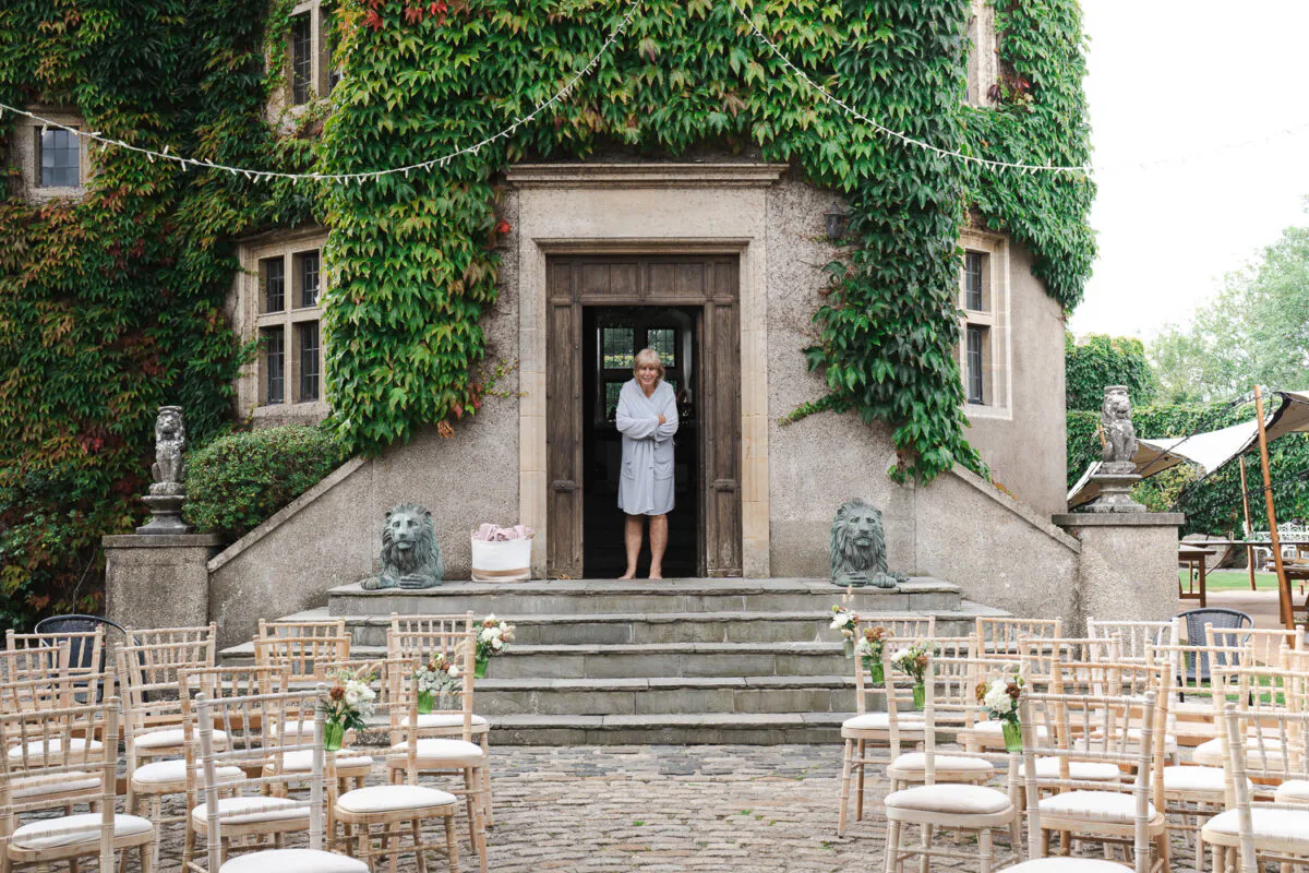 A mature bride stands smiling in her robe inside the wooden doorway of an ivy-covered stone castle, overlooking an outdoor ceremony setup with chiavari chairs.