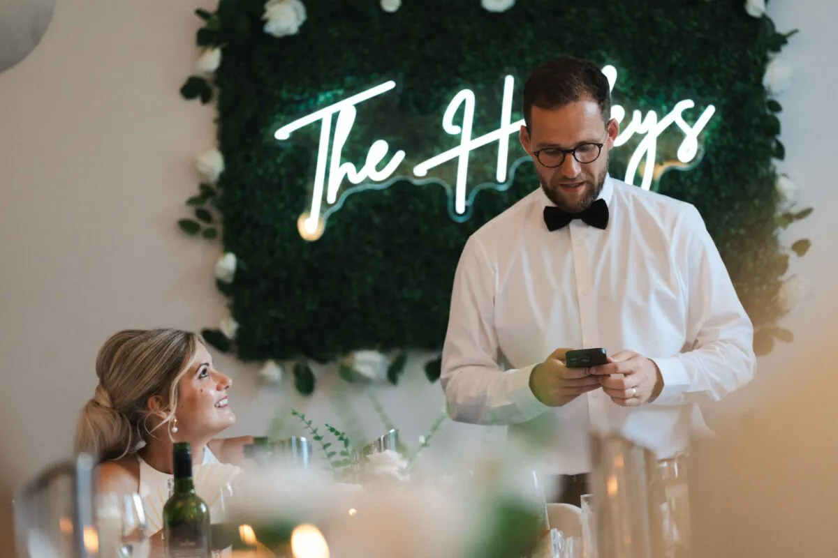 A groom in a white shirt and black bow tie looks down at his phone during speeches while the bride looks up at him smiling in front of a neon sign and greenery wall.