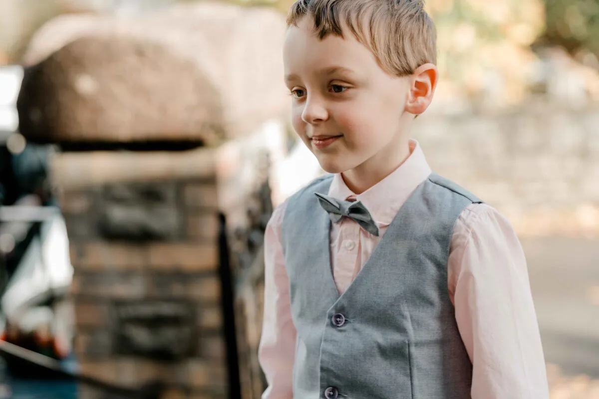 A young page boy wearing a grey waistcoat and bow tie smiles gently outdoors during a wedding.