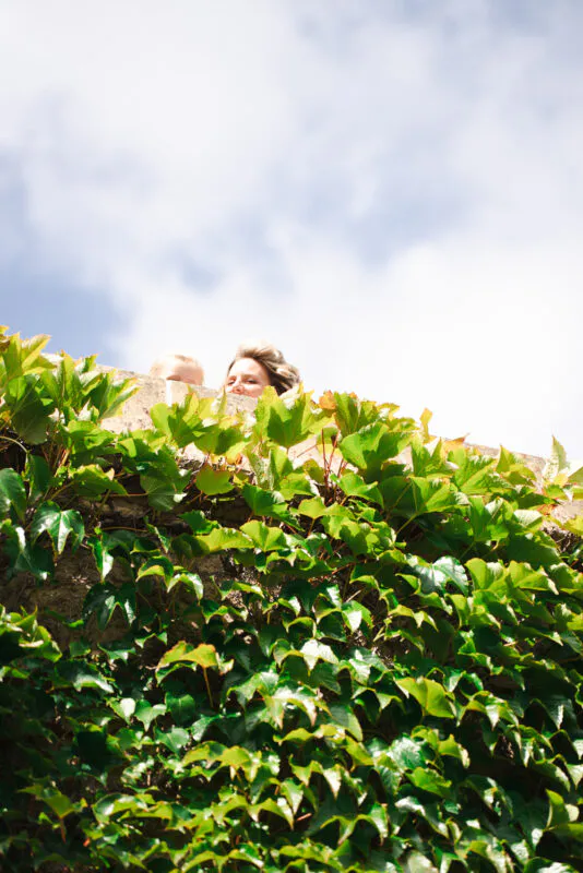 A low angle shot captures wedding guests peering over a stone wall thickly draped with vibrant green ivy beneath a bright cloudy sky.