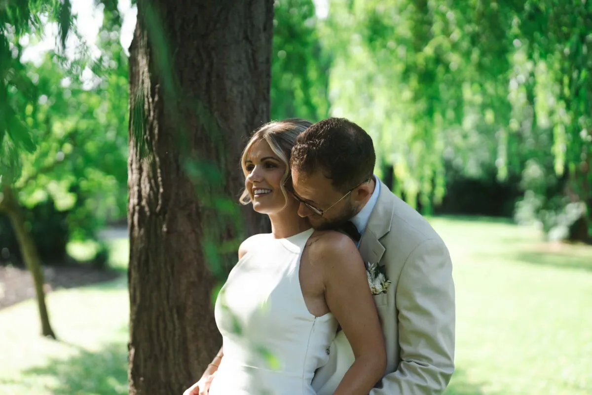 A smiling bride in a modern halter-neck gown stands outdoors in a woodland setting while her groom in a beige suit gently nuzzles her neck.