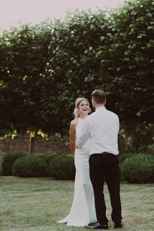 A newly married couple share a joyful laugh together outdoors in a manicured garden setting during their wedding reception.