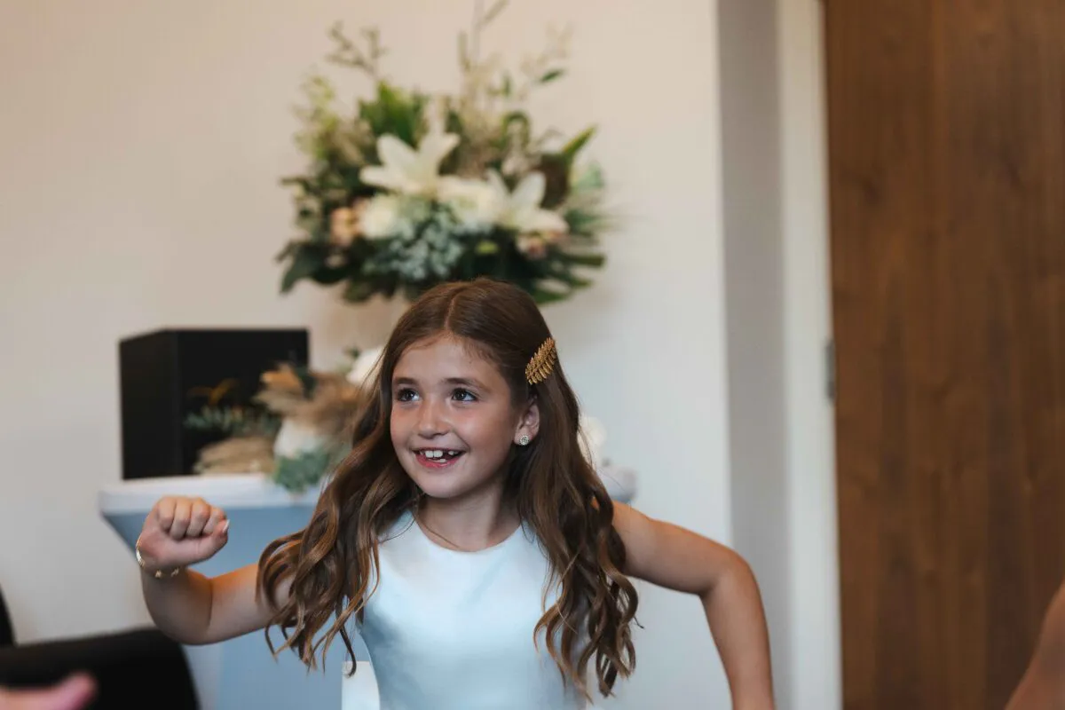 A young flower girl laughs joyfully with long wavy hair and a gold hair accessory while celebrating at a wedding reception.