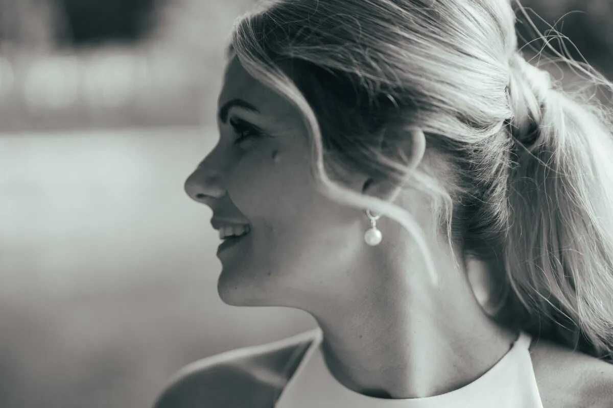 A close-up profile portrait in black and white capturing a smiling bride with a high ponytail and pearl earrings.