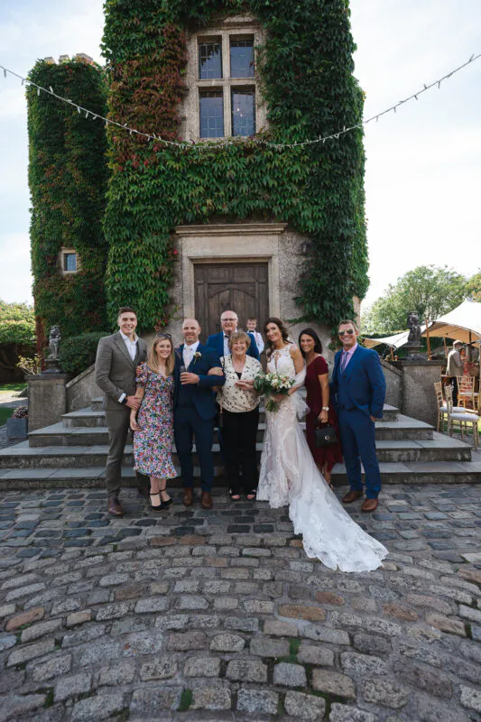 A bridal party and family members pose for a group photo outside a historic castle tower covered in lush green ivy.
