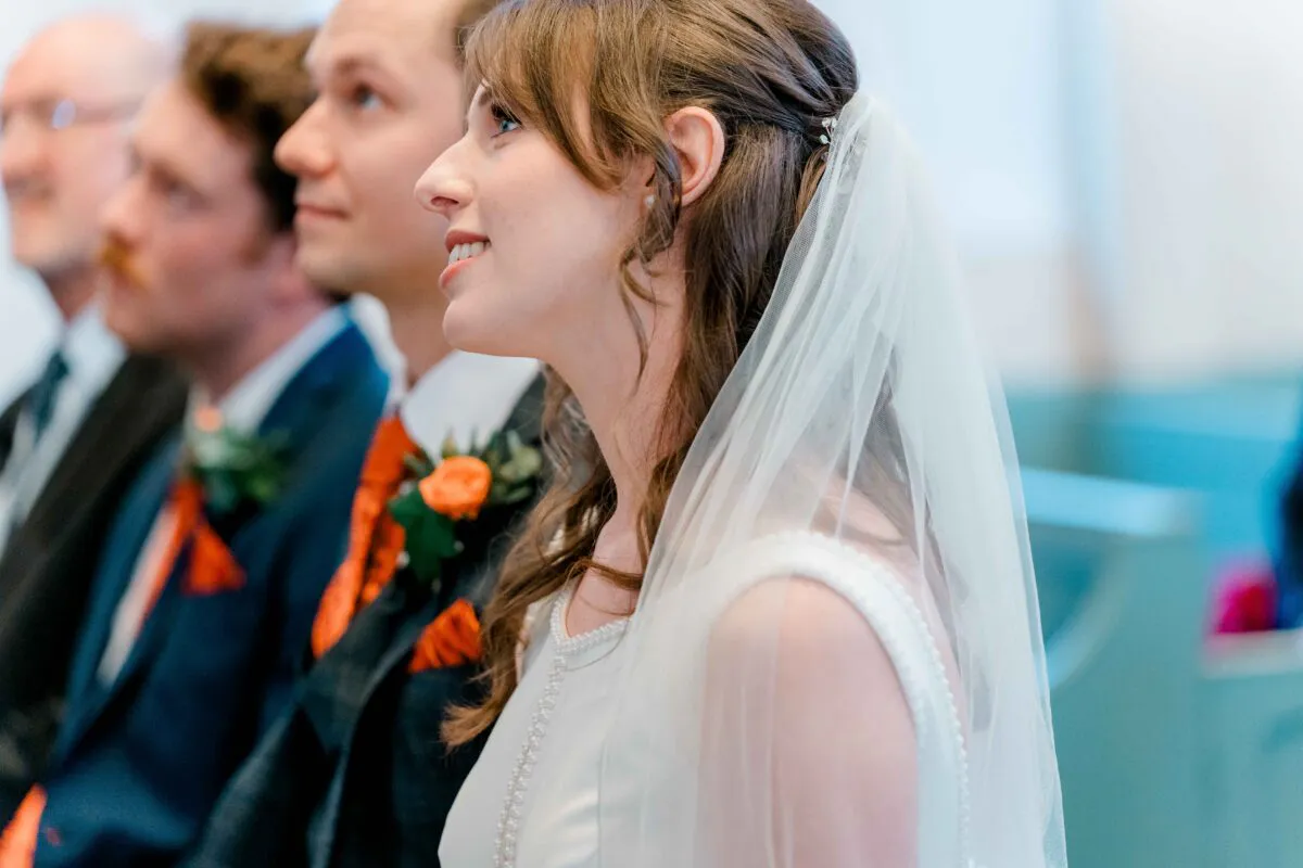 A side profile of a smiling bride wearing a half-up hairstyle and veil, seated alongside her groom and guests during a church ceremony.