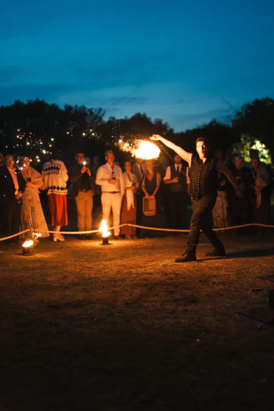 A fire performer entertains wedding guests gathered outdoors under a deep blue evening sky during the reception celebrations.