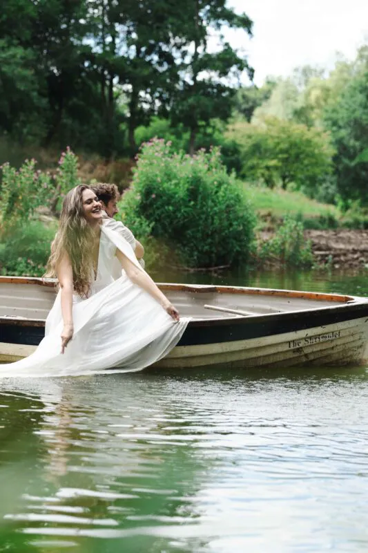 A newlywed couple laugh joyfully together while sitting inside a wooden rowing boat named The Shrimplet on a calm lake surrounded by lush greenery.