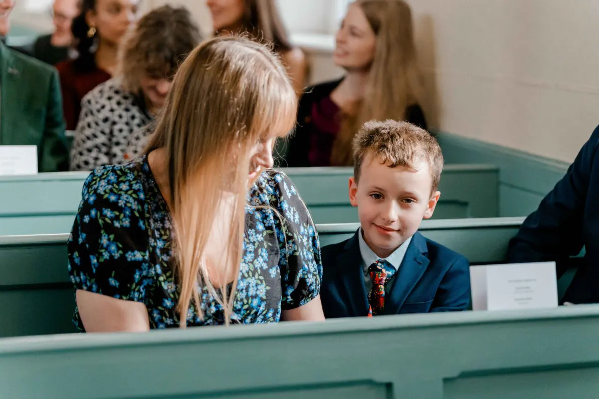 A young boy in a blue suit and patterned tie sits next to a woman in a floral dress on green wooden church pews during a wedding.