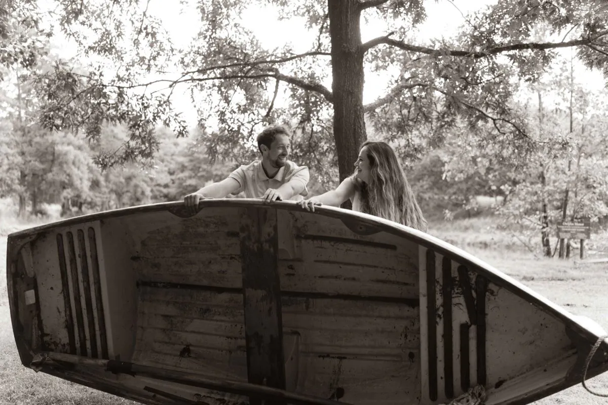A newlywed couple laughing and sharing a tender glance while leaning against an overturned wooden rowboat in a lush woodland setting.