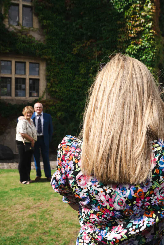 A wedding guest with long blonde hair and a floral dress takes a photo of an older couple posing outdoors in front of an ivy-covered stone building.