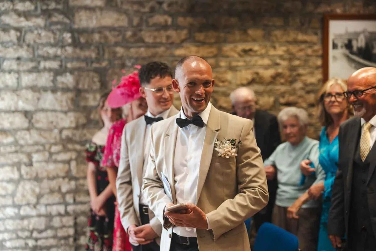A smiling groom in a beige suit and black bow tie stands in front of a rustic stone wall during a wedding ceremony.