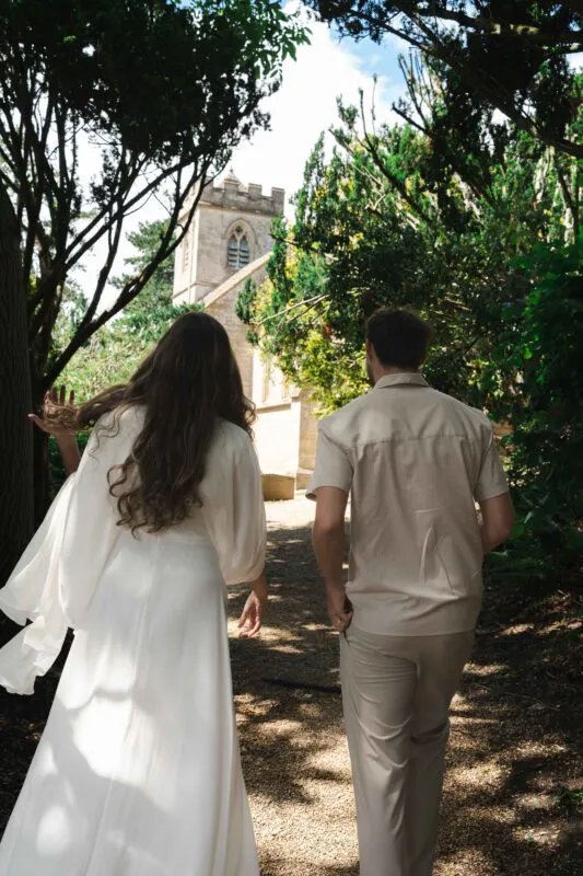 A newlywed couple walk away along a sunlit gravel path towards a historic stone church, framed by lush overhead trees.