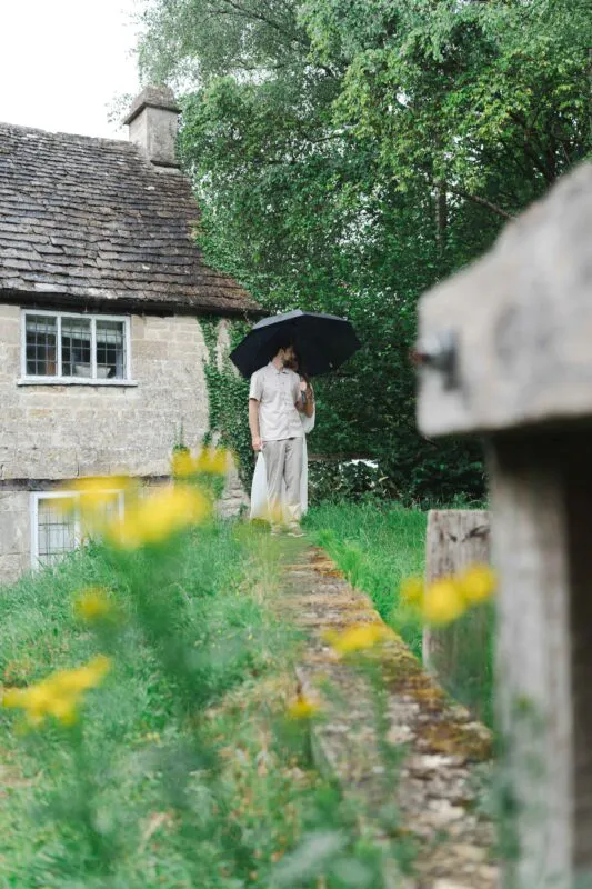 A newlywed couple stand together under a black umbrella on a rainy day in the garden next to a historic stone cottage.