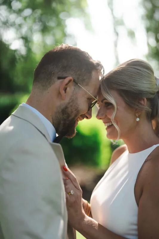 A newlywed couple share a tender forehead-to-forehead moment outdoors, bathed in warm backlit sunshine filtering through the trees.