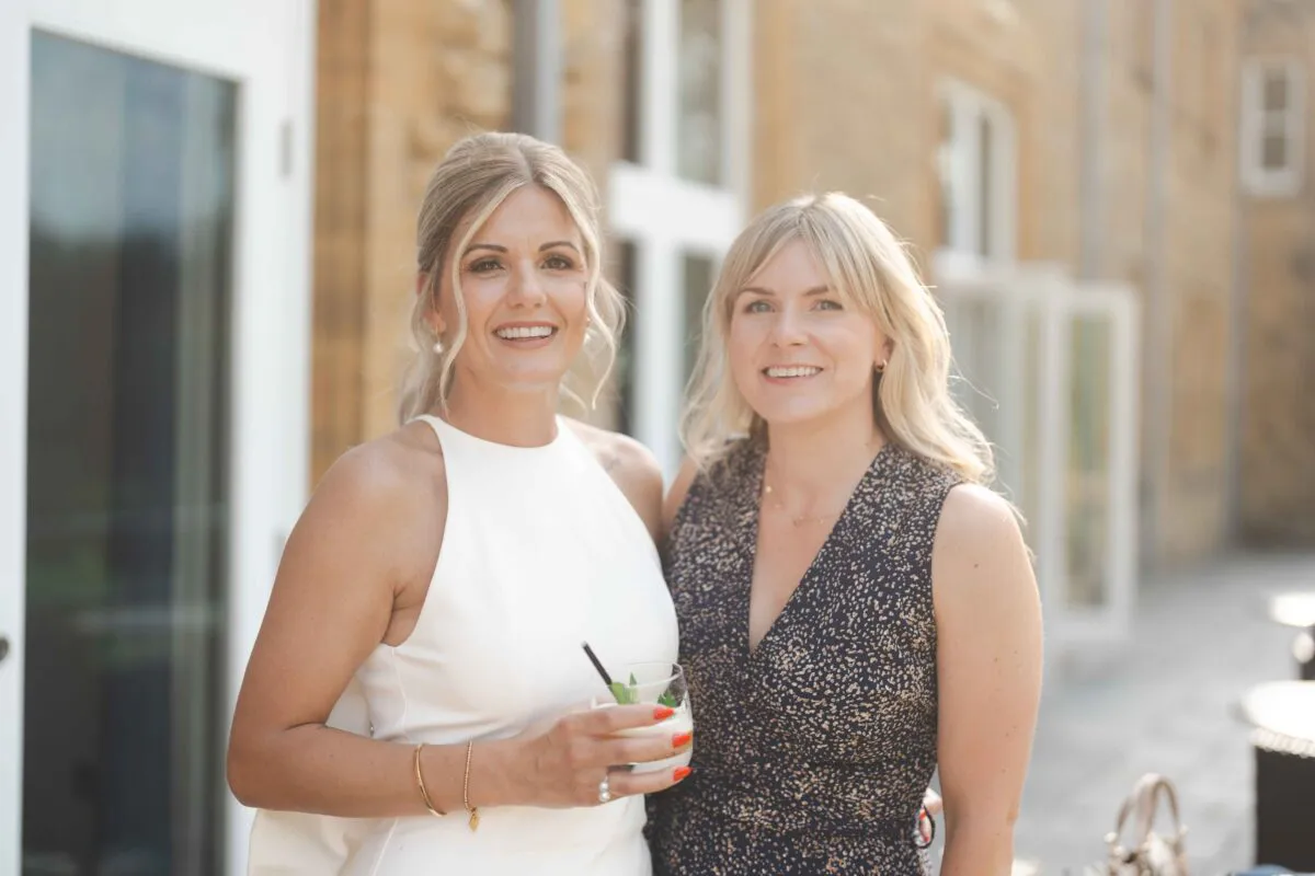 Two smiling female guests attending a daytime wedding reception outdoors while holding a cocktail glass.