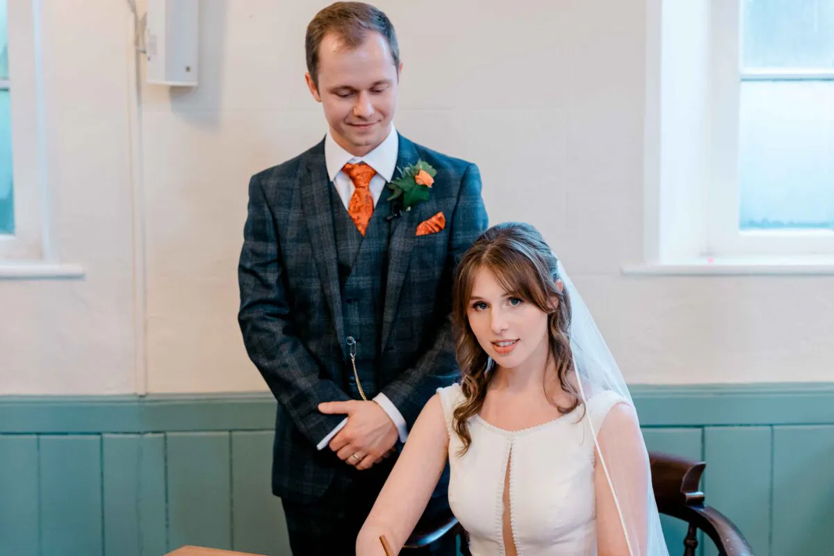 The bride sits signing the marriage register while the groom looks on smiling warmly, wearing a checked three-piece suit with an orange tie.