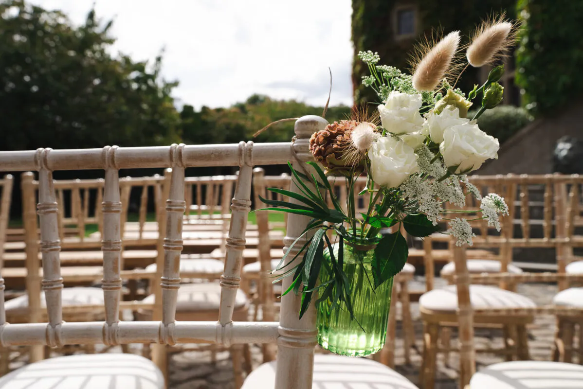 A rustic wedding chair is decorated with a small green glass bottle holding a floral arrangement of white roses and dried grasses for an outdoor ceremony.
