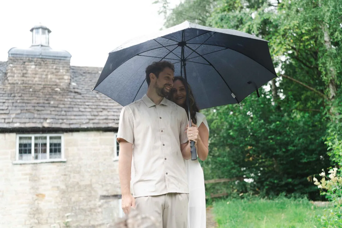 A newlywed couple smiling together under a black umbrella while standing outdoors in the rain near a stone building.
