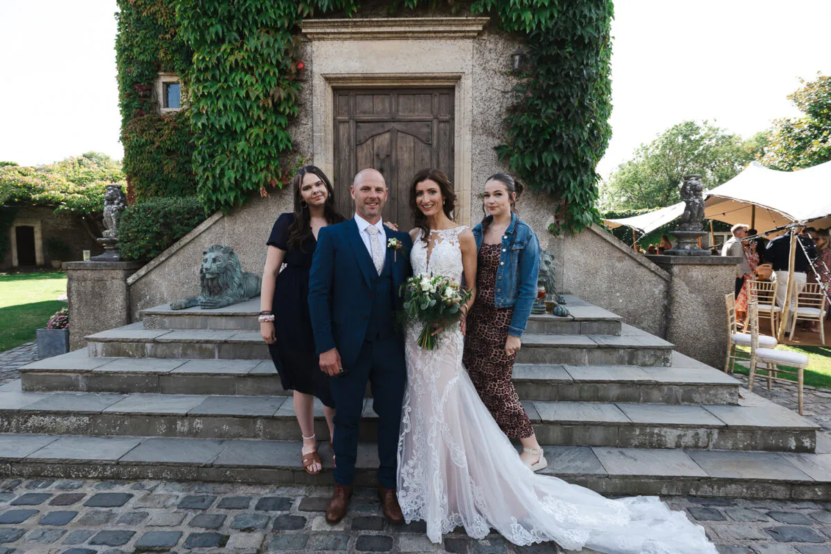 A newlywed couple posing with guests on stone steps outside an ivy-covered stone building on their wedding day.