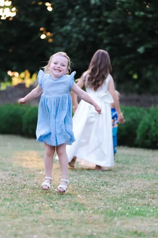 A young flower girl in a blue dress joyfully jumps on a lawn during an outdoor wedding reception while other children play in the background.