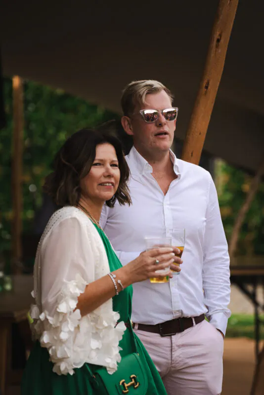 A smiling wedding guest holding a drink stands alongside a man wearing sunglasses during a relaxed drinks reception under a marquee.