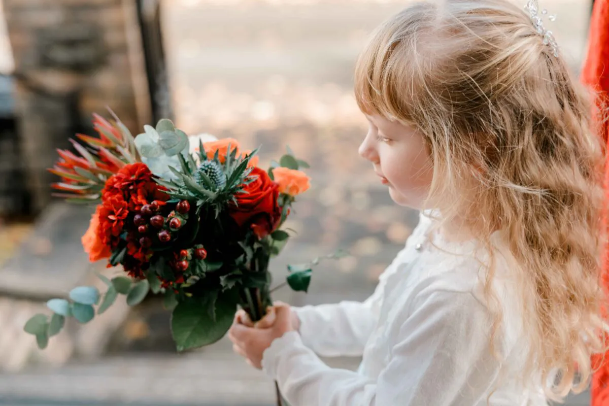 A young flower girl with long blonde hair holding a vibrant autumnal bouquet featuring red roses, thistles, and berries outdoors.